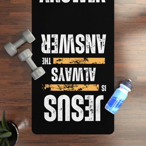 Puede incluir: Estera de yoga negra con texto blanco que dice "Jesús es siempre la respuesta". El texto tiene un aspecto desgastado. La estera está sobre un suelo de madera con una botella de agua y pesas.