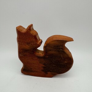 Könnte beinhalten: Eine Holzfigur eines Eichhörnchens, geschnitzt aus dunklem, braunem Holz. Das Eichhörnchen ist in sitzender Position dargestellt, mit nach oben gebogenem Schwanz. Die Holzmaserung ist sichtbar und verleiht dem handgefertigten Stück Textur. Die Figur steht vor einem schlichten, weißen Hintergrund.