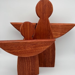 Pode incluir: Duas esculturas de anjos de madeira castanho-avermelhada. O anjo maior tem asas abertas e cabeça arredondada. O anjo menor está à frente, com um design semelhante. As esculturas estão sobre um fundo branco.