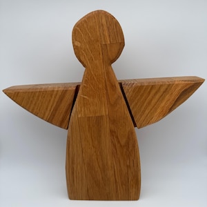 Pode incluir: Uma escultura de anjo de madeira com asas abertas. A escultura é feita de madeira castanha clara e tem um design simples e geométrico. As asas são retangulares e estendem-se do corpo. O anjo tem uma cabeça arredondada e um corpo afilado.
