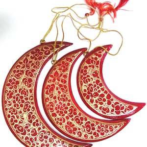 Peut inclure: Trois ornements en forme de croissant de lune rouge avec des motifs floraux dorés. Les ornements sont suspendus à des cordons dorés avec des glands rouges.