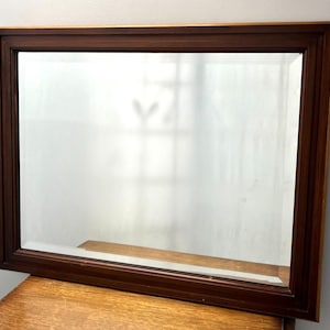 May include: A rectangular wall mirror featuring a dark brown wooden frame with a stepped, tiered profile. The glass surface is clear and reflective, showing a slight bevel along the inner edges where it meets the wood frame construction.