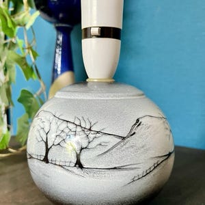 May include: A spherical ceramic lamp base featuring a textured light gray and white finish with a hand-painted black landscape design depicting bare trees and rolling hills. The top includes a white plastic light socket with a small brown switch.