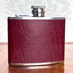 May include: A rectangular metal flask wrapped in a deep burgundy, textured faux leather material. The flask features a polished silver-tone metal frame along the top and bottom edges, with a matching silver-tone screw-top cap at the centre.