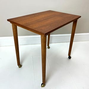 May include: A rectangular wooden side table with a smooth, medium-brown grain finish. The table features four tapered legs, each equipped with a small, dark-coloured castor wheel at the base, allowing for mobility across the floor surface.