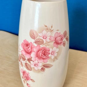 May include: A cylindrical ceramic vase with a smooth cream-colored finish. The exterior is decorated with a floral pattern featuring clusters of pink roses and smaller white blossoms, accompanied by light brown leaves and stems.