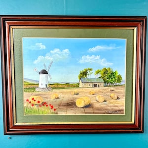 May include: A framed landscape painting depicting a white windmill and a stone building in a field with scattered hay bales under a blue sky with white clouds. Bright red poppies grow in the foreground, all enclosed in a dark wood frame.