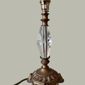 May include: A vintage table lamp featuring a clear, faceted crystal centre and a brass base. The base has an ornate, floral design. The lamp has a standard bulb socket and a brown electrical cord.