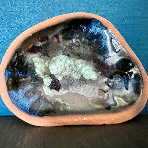 May include: An irregular, oval-shaped ceramic dish with a terracotta-colored rim. The interior features a glossy, multi-colored glaze with swirls of deep blue, black, and white, centered around a textured, pale mint-green crystalline formation.
