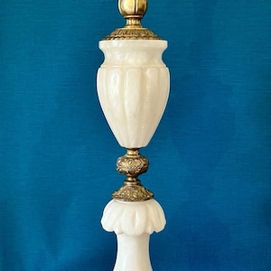 May include: A tall table lamp featuring a white milk glass body with two distinct sections, separated and topped by ornate brass metal components. The base is a wide, circular brass stand with intricate floral relief detailing and a smooth finish.