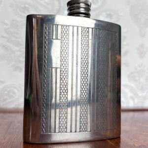 May include: A rectangular silver-toned metal flask featuring a textured front panel with alternating vertical stripes of smooth polished metal and a fine cross-hatched diamond pattern. The flask has a rounded top with a screw-on metal cap.