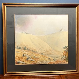 May include: A framed watercolor landscape painting depicting rolling hills in shades of beige and tan, with scattered rocks in the foreground, a small cluster of trees on the right, and a soft, hazy sky above, all enclosed in a gold-toned frame.