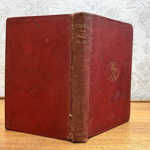 Peut inclure: Un livre ancien à couverture rigide rouge intitulé Through the Looking Glass. Le dos présente des lettres dorées et la couverture avant affiche un emblème circulaire doré. La couverture en tissu montre des signes d'usure et des bords effilochés.