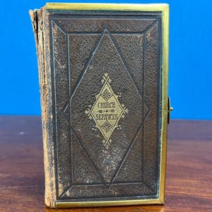 May include: A vintage book titled Church Services with a dark brown textured leather cover, featuring a central diamond-shaped gold embossed emblem and a brass metal frame border. The spine shows signs of wear and age with frayed edges.