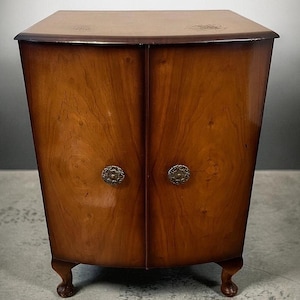 May include: A vintage wooden cabinet with a curved front and two doors. The cabinet is a warm brown colour with decorative metal knobs and rests on short, curved legs. The top of the cabinet is a darker shade of brown.