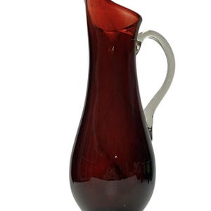 Könnte beinhalten: Eine hohe, elegante Glaskaraffe in tiefem Burgunderrot. Sie hat einen schlanken, länglichen Körper, einen ausgestellten Ausguss und einen Griff aus klarem Glas. Die Karaffe hat einen abgerundeten Boden und eine glatte, glänzende Oberfläche.