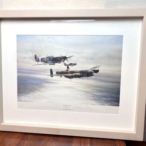 May include: A framed aviation art print titled 'Memphis Flight' featuring a dark-colored bomber aircraft flying in formation with two smaller fighter planes over a cloudy sky. The print is encased in a light-colored wooden frame with a white mat.