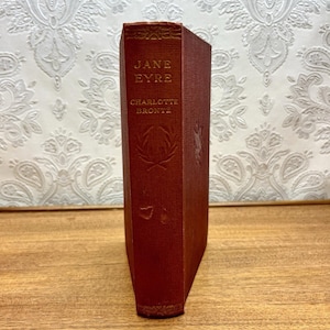 May include: A hardback book titled Jane Eyre by Charlotte Brontë with a textured, deep red fabric cover. The spine features gold-coloured embossed lettering and a decorative laurel wreath emblem centred below the author's name.