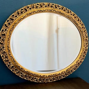 Può includere: Uno specchio da parete rotondo con un'ampia cornice dorata decorata, caratterizzata da intricati motivi a volute in rilievo e dettagli a forma di foglia. La superficie in vetro riflettente è liscia e chiara, inserita nel bordo metallico decorativo che circonda l'intera circonferenza dello specchio circolare.