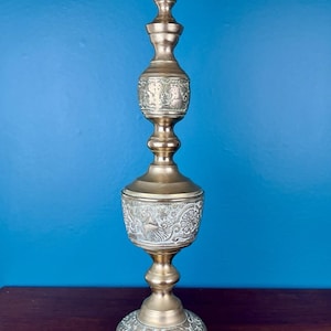 Puede incluir: Una base de lámpara de mesa alta y delgada de latón con un acabado metálico pulido. La estructura presenta un diseño ornamentado y escalonado con tres secciones distintas decoradas con intrincados patrones florales y vegetales en relieve.