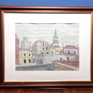 May include: A framed drawing of a European cityscape featuring stone buildings with red-tiled roofs and a central church spire. The artwork is rendered in muted tones of beige, grey, and red, enclosed in a dark wood frame with a gold inner trim.