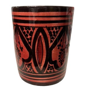 Può includere: Una tazza in ceramica con un design rosso e nero. La tazza ha una forma cilindrica con bordo e base marrone scuro. Il corpo rosso è decorato con motivi geometrici e floreali neri.