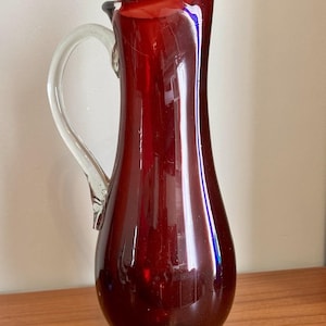 May include: A tall, deep red glass jug featuring a smooth, bulbous body that tapers towards a flared spout. The vessel includes a clear, curved glass handle attached to the side and rests on a circular, solid red glass base.