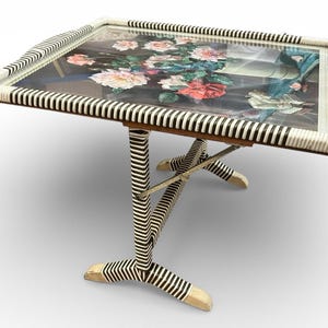 May include: A rectangular serving tray table with a floral design under glass. The frame and legs are white with black stripes. The legs are in an X-shape. The tray has handles on the sides.