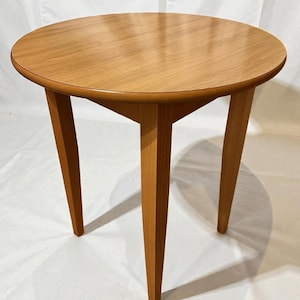 Può includere: Un tavolino rotondo in legno con tre gambe angolate. Il piano del tavolo è di colore marrone chiaro e le gambe sono di una tonalità leggermente più scura. Il tavolo ha un diametro di circa 61 cm e un'altezza di 51 cm.