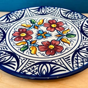 May include: A round ceramic plate featuring a white background with a central floral motif in shades of red, yellow, green, and blue. The border is decorated with intricate dark blue geometric and leaf patterns, creating a symmetrical design.