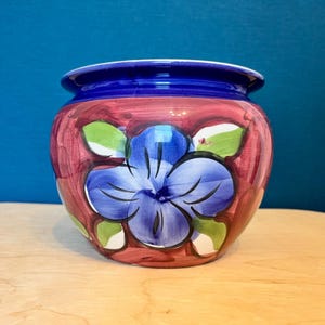 May include: A ceramic planter with a wide, rounded body and a thick, dark blue rim. The exterior features a hand-painted design of a large, central blue flower with green leaves, set against a background of blended, streaky reddish-pink brushstrokes.
