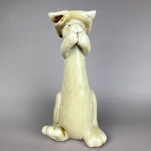 May include: A cream-colored ceramic figurine of a rabbit wearing a wide-brimmed hat. The rabbit is depicted in a seated position with its front paws raised to its face, covering its mouth. The glaze has a glossy finish with subtle brown accents.