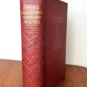 May include: A red hardcover book titled General Engineering Workshop Practice with gold lettering on the spine. The spine features horizontal lines and a repeating embossed pattern. The front cover has a faint, rectangular embossed design.