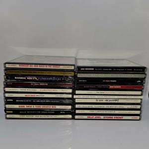 Może przedstawiać: Stos płyt kompaktowych (CD) z różnymi artystami i tytułami albumów. Płyty CD są ułożone pionowo, z tytułami skierowanymi do przodu. Niektórzy z artystów to Midnight Oil, Bonnie Raitt, Hank Williams, Miles Davis, Brooks & Dunn, Blood, Sweat & Tears, Willie Nelson, Toby Keith, The Fixx, Roy Orbison, Alison Krauss, Tina Turner, Zac Brown Band, The J. Geils Band, UB40 i Billy Joel.