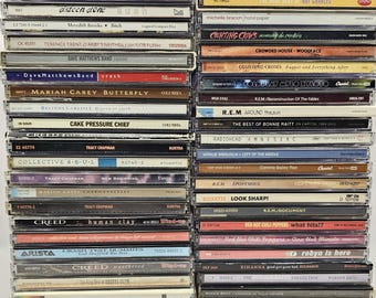 Puede incluir: Dos pilas verticales de CD de música en cajas de plástico. Los lomos muestran varios nombres de artistas y títulos de álbumes, incluidos Gavin DeGraw, Mariah Carey, Creed, R.E.M., Red Hot Chili Peppers y Bonnie Raitt, en una cuadrícula.