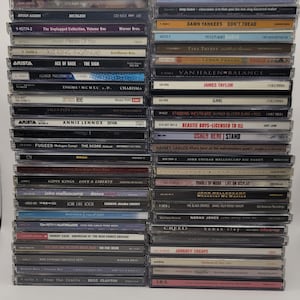 Puede incluir: Una pila de discos compactos (CD) con varios artistas y álbumes. Los CD están dispuestos verticalmente, con las espinas hacia el espectador. Algunos de los artistas que aparecen son: Inner Circle, Bryan Adams, Jewel, kd lang, Ace of Base, Ennio Morricone, Annie Lennox, Fugees, Gipsy Kings, John Mellencamp, Madonna, Tom Petty, Johnny Cash, Bob Seger, Rod Stewart, Eric Clapton, Damn Yankees, Michael Bublé, Tina Turner, Van Halen, James Taylor, Stabbing Westward, Beastie Boys, Usher, Harvey Danger, John Cougar Mellencamp, Puddle of Mudd, The Black Crowes, Norah Jones, Creed, Journey, I.R.S.