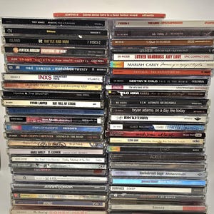 Op de afbeelding: Een grote verzameling muziek-cd's gestapeld in twee verticale kolommen. De ruggen tonen verschillende artiestennamen en albumtitels, waaronder Jason Mraz, U2, Mariah Carey, Destiny's Child, Adele en Justin Bieber, met zichtbare catalogusnummers.