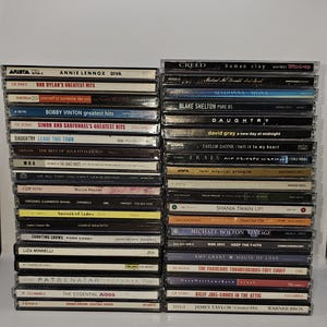 Może przedstawiać: Stos 20 płyt kompaktowych (CD) z różnymi artystami i albumami. Płyty CD są ułożone pionowo, z grzbietami zwróconymi do widza. Niektórzy z przedstawionych artystów to Bob Dylan, Simon and Garfunkel, Ella Fitzgerald, Creedence Clearwater Revival, Shania Twain, Michael Bolton, Bon Jovi, Amy Grant, Pat Benatar, The Judds, Billy Joel i James Taylor.