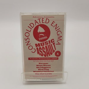 Op de afbeelding: Een doorzichtige cassetteband met een bedrukte inlay. De inlay toont de tekst "Consolidated Enigma Music Assault" in rood en wit, met een rood cirkelvormig logo. De tekst "August/September 1990" is ook zichtbaar.