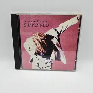May include: A CD case for the album "A New Flame" by Simply Red. The cover art depicts a person in a suit with arms raised against a pink backdrop. The album title is written in white text.