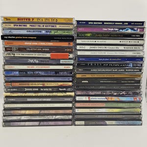 May include: Two vertical stacks of music CDs in plastic jewel cases. The spines display various artist names and album titles including Sister 7, Spin Doctors, Collective Soul, Ray Charles, Sheryl Crow, Tori Amos, 10,000 Maniacs, and Alanis Morissette.
