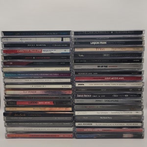 Op de afbeelding: Een stapel compact discs (cd's) met verschillende artiesten en albumtitels. De cd's zijn verticaal gerangschikt, met de ruggen naar de kijker gericht. Sommige van de artiesten zijn Christina Aguilera, Ricky Martin, Seal, Train en Janet Jackson.