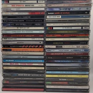 May include: A stack of compact discs featuring various artists and albums, including U2, Bruce Springsteen, Sting, and Fleetwood Mac. The CDs are arranged vertically, with the spines facing the viewer. The CDs are a mix of different colours and designs.