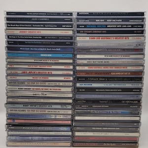 May include: A stack of compact discs featuring various artists and albums, including Journey, Janis Joplin, Bryan Adams, Elton John, and The Eagles. The discs are arranged in a vertical stack, with the titles and artist names visible on the spines.