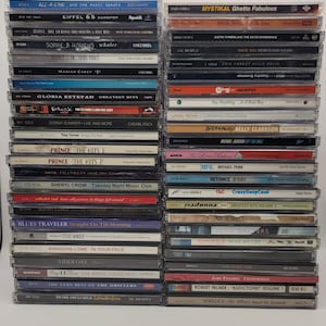 May include: A stack of compact discs (CDs) with various artists and album titles. The CDs are arranged vertically, with the spines facing the viewer. Some of the artists featured include Mariah Carey, Gloria Estefan, Prince, and Madonna.