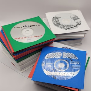 May include: Stacks of CDs in colorful sleeves. One CD is labeled "tracy chapman matters of the heart" in a green sleeve. Another CD is labeled "Snoop Dogg" in a blue sleeve. A third CD is in a white sleeve.