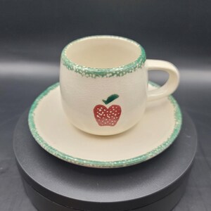 May include: A white ceramic teacup and saucer set with a green rim and a red apple design.