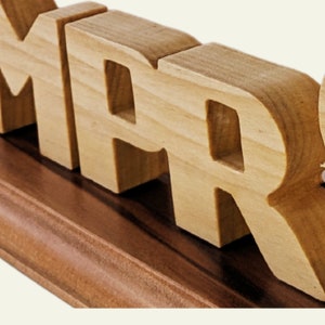 May include: Wooden MAR letters, carved from light-colored wood, are mounted on a dark brown wooden base. The letters are thick and blocky, with a natural wood grain visible. The base has a contrasting, darker finish.