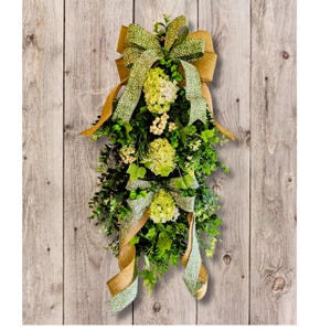 May include: A green and gold swag with a large bow and white flowers. The swag is hanging on a wood background.