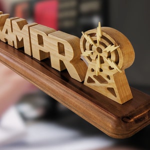 May include: Wooden desk sign with the name "KAMAR" in block letters, accompanied by a radio tower design. The sign is made of light-colored wood and mounted on a dark brown base. The radio tower has a circular design with lightning bolt accents.